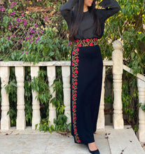 Person wearing a black outfit with floral embroidery standing on a stone path with greenery in the background